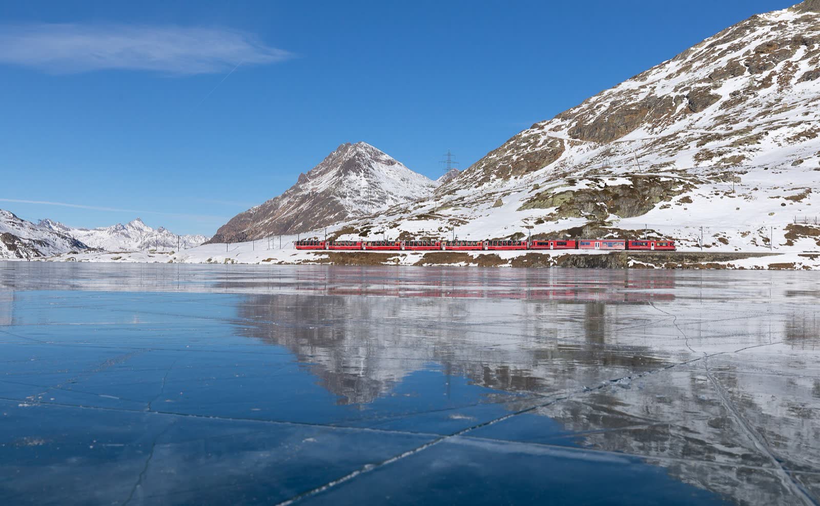 El Bernina Express rojo bordeando el Lago Bianco, con las montañas nevadas reflejadas en el agua